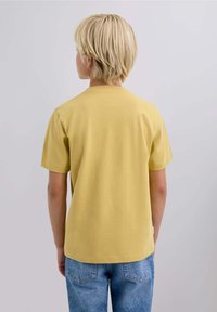 Kortærmet gul bomulds t-shirt med rund hals, båret med blå denimjeans. Stoffet har en glat, blød tekstur.