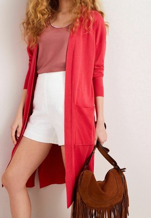 Femme portant un long cardigan rouge, un top mauve, un short blanc, tenant un sac à main marron à franges à côté d’elle.