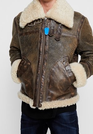 Braune, abgenutzte Leder-Shearlingjacke mit großem cremefarbenem Pelzkragen, -manschetten und -saum, getragen über dunkler Jeans, Hände in den Taschen.