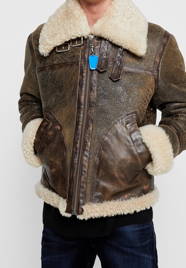 Veste en cuir marron usée avec doublure en shearling, grand col en fourrure crème, poignets et ourlet, portée sur un jean foncé avec les mains dans les poches.