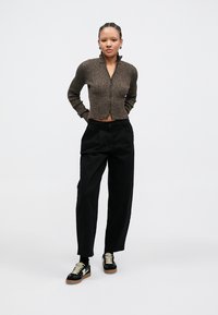 Brown ribbed cropped cardigan, black loose-fit trousers, and black sneakers with cream laces. Model stands with hands on hips against a plain background.