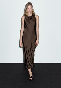 Brun ärmlös klänning med en slät, glänsande textur, prydd med ett runt, mörkt pärlhalsband och svarta sandaler. Modellen står framför en vit bakgrund.