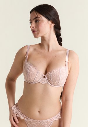 Donna con capelli lunghi e scuri raccolti in una treccia, indossa un reggiseno di pizzo rosa chiaro e mutandine di pizzo abbinate, guarda verso sinistra su uno sfondo chiaro.