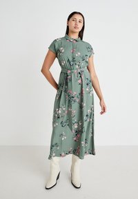 Robe midi à imprimé floral vert avec des manches courtes, taille ceinturée et boutonnage sur le devant. Présente des détails de fleurs roses et bleues.