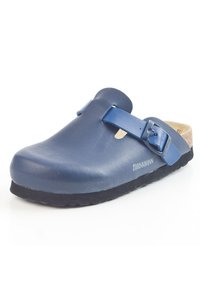 Zoccolo in pelle blu con punta arrotondata, cinturino regolabile e soletta in sughero. Suola in gomma nera texturizzata con un dettaglio di branding discreto.