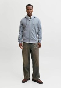 Sweat à capuche gris clair zippé avec une capuche à cordon, accompagné d'une chemise à carreaux. Pantalon en denim foncé délavé coupe décontractée et chaussures marron.