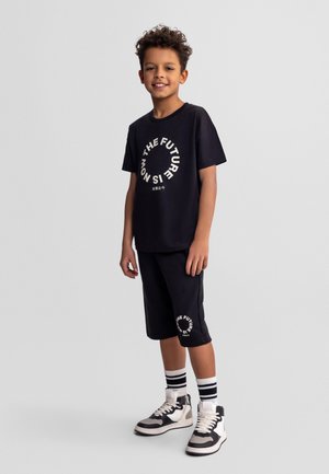 Chłopiec z kręconymi włosami, ubrany w czarne spodenki i koszulkę z napisem "Przyszłość jest teraz", białe i czarne trampki oraz paskowane skarpetki, stoi i się uśmiecha.