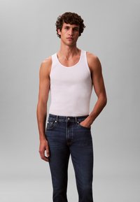 Balts kokvilnas topiņš bez piedurknēm un tumši zili džinsa džinsi. Topiņš ir piegulošs ar platām plecu siksnām, bet džinsiem ir klasiskā piecu kabatu dizains.