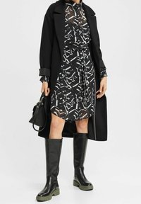 Schwarzer langer Mantel über einem gemusterten schwarz-weißen Kleid mit tailliertem Bund. Knielange schwarze Stiefel mit grünen Sohlen. Kleine schwarze Handtasche.