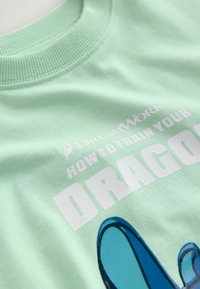 Mintgrön bomullströja med rundad halsringning och tryckt text "DreamWorks HUR MAN TRÄNAR DIN DRAGON" tillsammans med en blå drakgrafik.