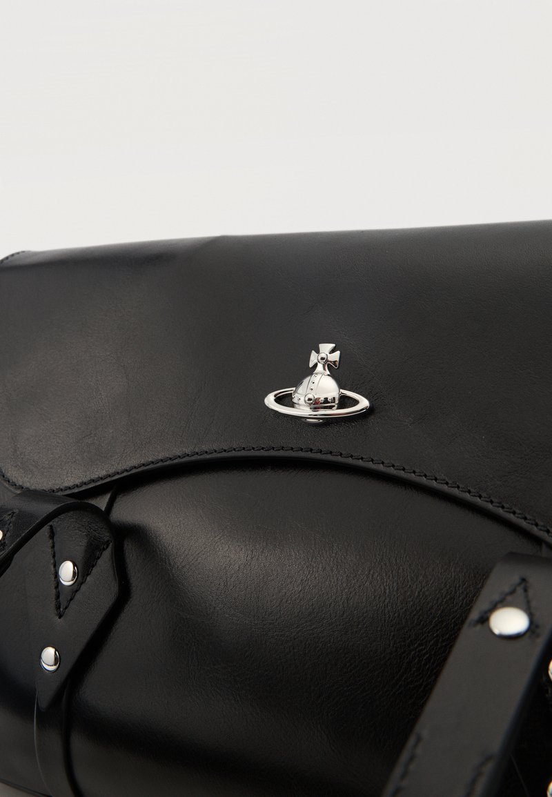 Sac en cuir noir avec un emblème sphérique argenté présentant une croix et un détail d'anneau sur le rabat, avec des coutures visibles et des sangles.