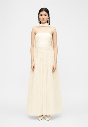 Mujer con vestido crema sin tirantes hasta el suelo con bordado floral, combinado con pañuelo a juego y pendientes de perlas colgantes, de pie inmóvil.