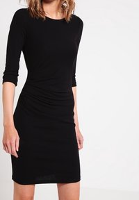 Robe noire à manches longues avec une silhouette ajustée et un détail froncé sur le côté, en tissu doux et extensible.