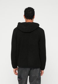 Musta fleece-takki hupulla, rento istuvuus, teksturoitu pinta ja sauma hartioiden yli. Yhdistetty tummanharmaisiin housuihin.