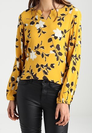 Femme portant un chemisier jaune à fleurs à manches longues avec un col en V et un pantalon en cuir noir ajusté.