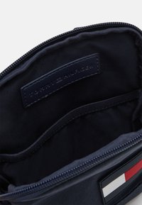 Otevřená námořnická modrá zipová taška, která ukazuje vnitřní štítek s textem "Tommy Hilfiger" a částečný pohled na červenobílo-modrou logo značky.