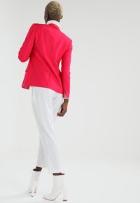 Jasně růžové sako na míru, bílé kalhoty s tenkými proužky a bílé kotníkové boty na vysokém podpatku. Hladká textura, elegantní design.