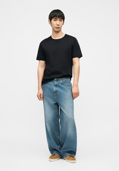 Νεαρός άνδρας που στέκεται φορώντας μαύρο κοντομάνικο μπλουζάκι, φαρδιά ανοιχτό μπλε τζιν και καφέ αθλητικά παπούτσια σε απλό λευκό φόντο.