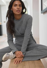 Grå, langærmet jumpsuit i blødt materiale med omslagsdesign og sidelukninger, parret med sølv sneakers. Model siddende på en træoverflade.