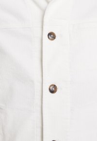 White corduroy shirt featuring vertical ribbing, five brown buttons, and a structured collar design. Suitable for casual wear.