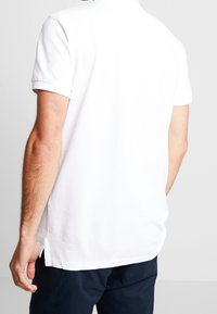 Vit polo-tröja med korta ärmar, med en klassisk krage, sidokilar och en mjuk bomullstextur. Bärs med marinblå byxor.