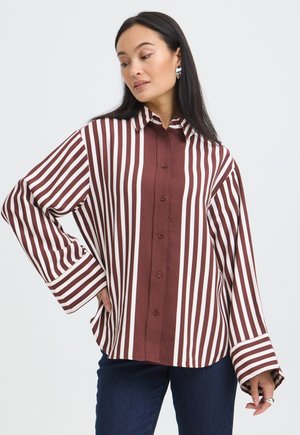 Femme aux longs cheveux foncés portant une chemise ample à rayures verticales marron et blanches et un jean bleu, regardant sur le côté.