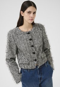 Jakna s teksturom u bijeloj i crnoj boji u uzorak houndstooth s izrobnim rubovima, okruglim izrezom, šest crnih dugmadi i prednjim džepovima. U kombinaciji s plavim trapericama.