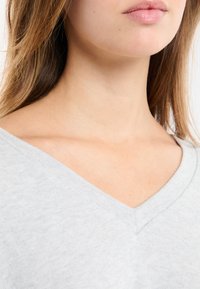 Szary sweter z dekoltem w serek, wykonany z miękkiej, teksturowanej tkaniny. Dekolt ma schludne wykończenie z subtelnym przeszyciem przy krawędzi.