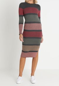 Femme portant une robe en maille rayée côtelée à manches longues et fente latérale, assortie à des baskets blanches, se tenant devant un fond uni.