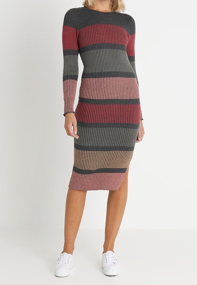 Femme portant une robe en maille rayée côtelée à manches longues et fente latérale, assortie à des baskets blanches, se tenant devant un fond uni.