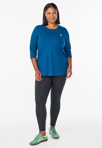 Blauwe sportieve lange mouw top met ronde hals, gecombineerd met zwarte leggings en groene sneakers; gladde textuur en casual ontwerp.