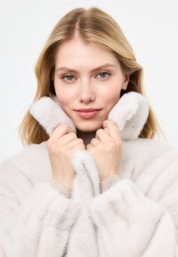 Cappotto in pelliccia sintetica grigio chiaro con colletto alto, polsini a coste e dettaglio decorativo con zip. Il modello tiene delicatamente il colletto vicino al viso.