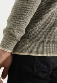 Maglione lavorato a maglia in tonalità marrone terroso con polsini e orlo a coste; presenta una texture intrecciata e un piccolo etichetta del logo sul lato.