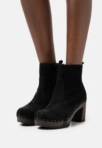 Botas de tobillo de ante negro con tacones de bloque y cremalleras laterales, lucidas en piernas de piel oscura sobre un fondo blanco liso.