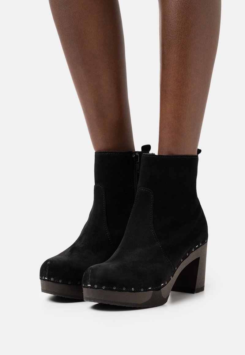 Botas de tobillo de ante negro con tacones de bloque y cremalleras laterales, lucidas en piernas de piel oscura sobre un fondo blanco liso.