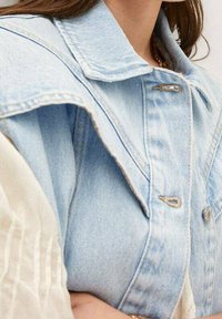 Helles blaues Denim-Weste mit einem auffälligen Kragen, silbernen Beschlägen und einem leicht übergroßen Design mit markanten Steppdetails.
