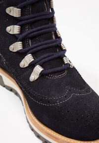 Bottine en suède bleu foncé avec ferrures métalliques et lacets, ornée de perforations décoratives et d'une semelle claire contrastante.