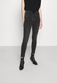 Černé džíny s vysokým pasem a zapínáním na knoflíky, dvěma předními kapsami a přiléhavým střihem. Kombinováno s černými kotníkovými botami a topem bez rukávů.