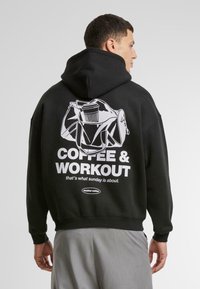 Zwarte sweatshirt met een relaxte pasvorm, grote capuchon en een grafisch ontwerp op de achterkant dat een sporttas, koffiekop en de tekst "KOFFIE & TRAINEN" afbeeldt.