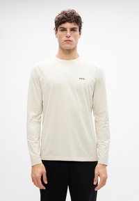 Tricou crem cu maneci lungi, fabricat dintr-un material moale, cu decolteu rotund, având un mic logo "BOSS" imprimat în negru pe piept.