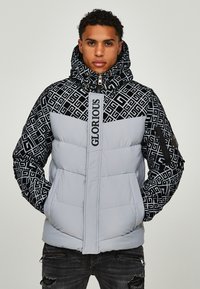 Giubbotto puffer grigio con cappuccio e maniche nere a motivo, dotato di logo "GLORIOUS" e tasche con zip. Design testurizzato e isolato.