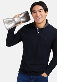 Un homme souriant portant un polo marine et un jean tient un grand nœud papillon en métal près de sa tête d'une main, l'autre main dans la poche.