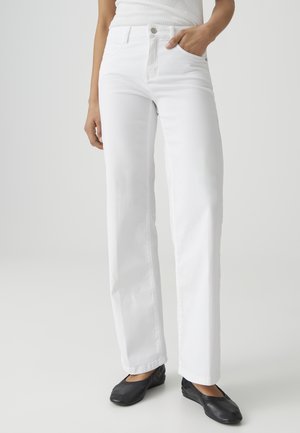 Frau trägt gerade geschnittene weiße Jeans und schwarze Ballerinas, mit einer Hand in der Tasche, vor einem einfarbigen hellen Hintergrund.