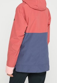 Wodoodporna kurtka z kapturem, z różową górną częścią i niebieską dolną częścią. Wykonana z trwałej tkaniny o gładkiej teksturze.