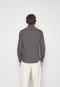 Man met middellang bruin haar, gekleed in een donkergrijze langemouwenshirt en lichtbeige broek, met zijn gezicht van de kijker af tegen een effen achtergrond.
