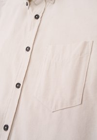 Beige button-up shirt made of textured fabric, featuring a chest pocket and dark buttons, with a classic collar design.