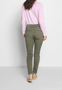 Oliwkowe wąskie dżinsy z tylnymi kieszeniami, przeszyciami i wysokim stanem, zestawione z jasnoróżową bluzką z długim rękawem oraz beżowymi sandałami.