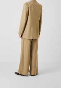 Béžový saka na míru s jedním rozparkem na zadní straně, zkombinovaný s kalhotami se širokými nohavicemi. Sako má čtyři knoflíkové detaily na rukávech.