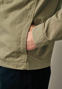Jachetă khaki cu o textură netedă, având o buzunar lateral și detalii la manșete. Designul este simplu și ușor, potrivit pentru îmbrăcăminte casual.