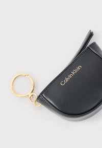 Μαύρο δερμάτινο πορτοφόλι κλειδιών Calvin Klein με χρυσό κρίκο κλειδιών και φερμουάρ σε λευκό φόντο.